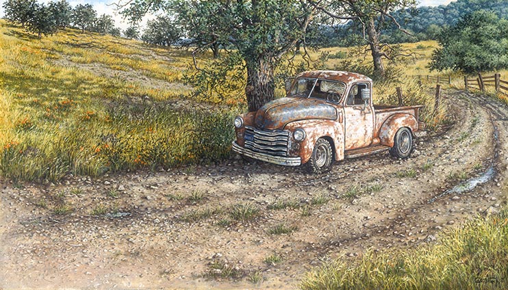 A rusted old truck sits where it was abandoned, along a dirt road next to a gnarled old tree. Recent rains have left thin little puddles along the wheel tracks in the road. Bright yellow wildflowers bloom in the fields surrounding the truck.